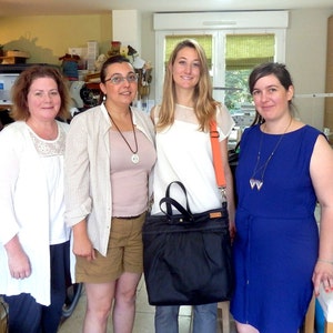 May include: Four women standing in a room, three are wearing casual clothing and one is wearing a blue dress. The woman in the center is holding a black bag with a zipper.