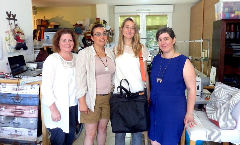 May include: Four women standing in a room, three are wearing casual clothing and one is wearing a blue dress. The woman in the center is holding a black bag with a zipper.