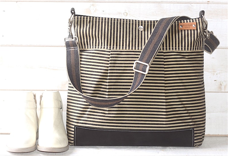 May include: A striped black and beige canvas tote bag with a brown leather strap and a brown leather bottom. The bag has a metal clasp and a small brown leather tag with the text "GRADAGE GOODS" on it.