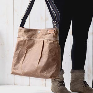 May include: A tan waxed canvas shoulder bag with a large main compartment, a top snap closure, and a dark blue and tan striped adjustable shoulder strap. A small rectangular leather brand tag is attached to the upper right corner of the bag.