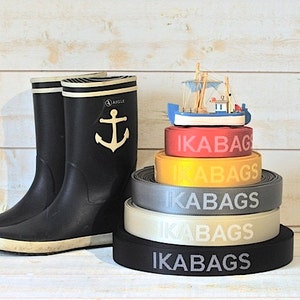 May include: A stack of five black, gray, beige, yellow, and red elastic straps with the text "IKABAGS" printed in white on each strap. A small wooden toy boat sits on top of the straps.