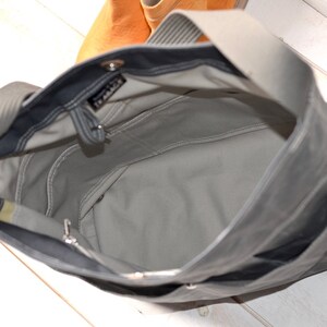 May include: A dark gray tote bag with a light gray interior lining, featuring a wide shoulder strap, an open top, and internal pockets. A small black label with white text reading "TE ABACK" is visible near the top edge of the bag interior.