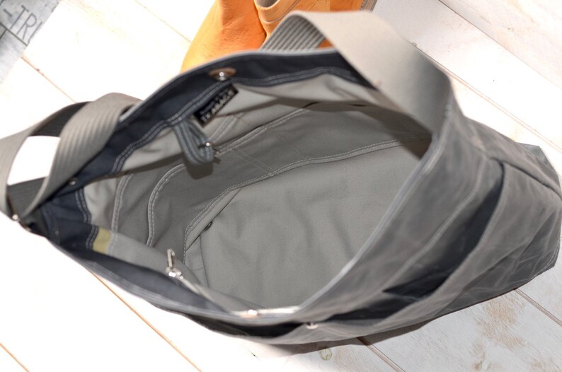 May include: A dark gray tote bag with a light gray interior lining, featuring a wide shoulder strap, an open top, and internal pockets. A small black label with white text reading "TE ABACK" is visible near the top edge of the bag interior.