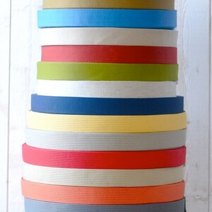 May include: A stack of 15 different colored cotton webbing straps. The straps are all the same width and are stacked on top of each other. The colors of the straps are gray, brown, blue, white, red, green, white, navy blue, yellow, gray, red, beige, orange, gray, and white.