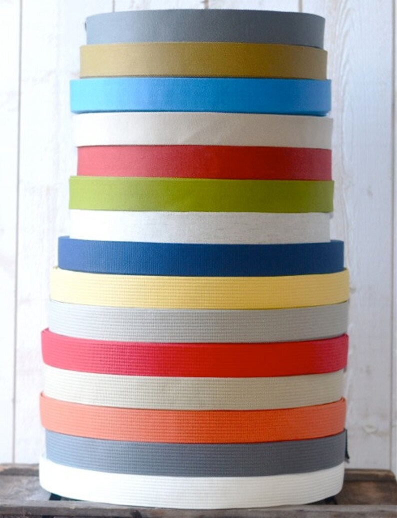 May include: A stack of 15 different colored cotton webbing straps. The straps are all the same width and are stacked on top of each other. The colors of the straps are gray, brown, blue, white, red, green, white, navy blue, yellow, gray, red, beige, orange, gray, and white.
