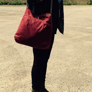 May include: A large, deep red shoulder bag with a slouchy, unstructured silhouette. The bag features a wide strap, a top opening, and a small rectangular leather-like patch detail near the upper corner, providing a simple and functional design.