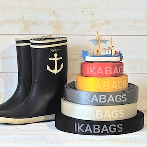 May include: A stack of five black, gray, beige, yellow, and red elastic bands with the word "IKABAGS" printed in white on each band. A small wooden toy boat sits on top of the stack of bands.