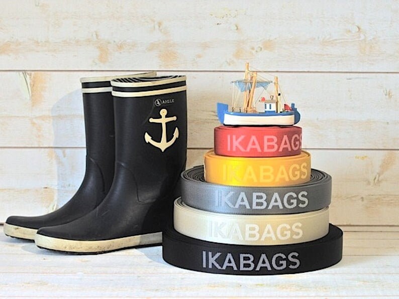 May include: A stack of five black, gray, beige, yellow, and red elastic bands with the word "IKABAGS" printed in white on each band. A small wooden toy boat sits on top of the stack of bands.