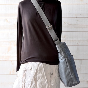 May include: A dark gray crossbody bag with a wide, adjustable gray strap and visible white stitching, worn over a black long-sleeved turtleneck shirt and a light beige skirt. The bag features a structured design with an external side pocket.