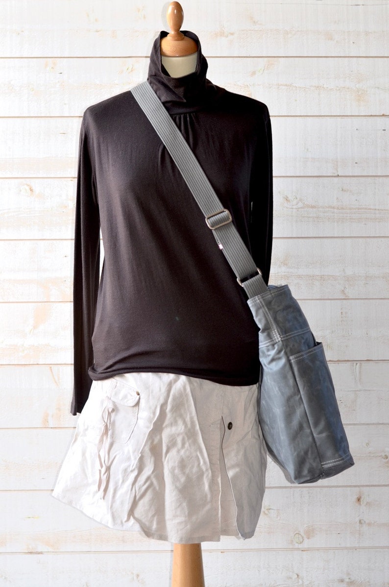 May include: A dark gray crossbody bag with a wide, adjustable gray strap and visible white stitching, worn over a black long-sleeved turtleneck shirt and a light beige skirt. The bag features a structured design with an external side pocket.