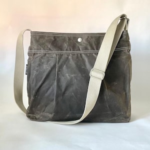 May include: A dark gray crossbody bag with a weathered texture, featuring a wide beige adjustable shoulder strap, white contrast stitching along the top panel, and a single silver-toned metal snap button closure at the center of the top edge.