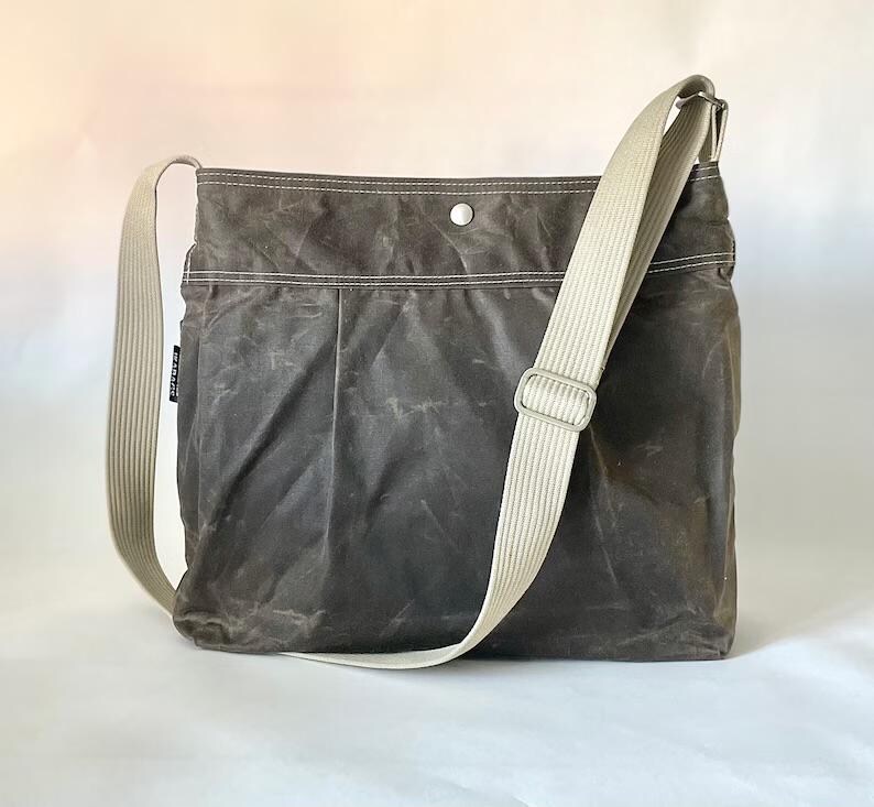 May include: A dark gray crossbody bag with a weathered texture, featuring a wide beige adjustable shoulder strap, white contrast stitching along the top panel, and a single silver-toned metal snap button closure at the center of the top edge.