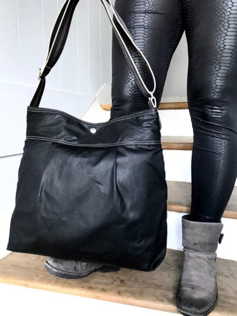 May include: A large black shoulder bag featuring a slouchy silhouette, a single silver-tone snap button closure at the top center, and a long adjustable strap with white trim detailing. The bag has visible white stitching along the top edge.