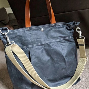 May include: A dark blue canvas tote bag with brown leather handles and a beige adjustable shoulder strap. The bag has a zipper closure and a silver metal clasp.