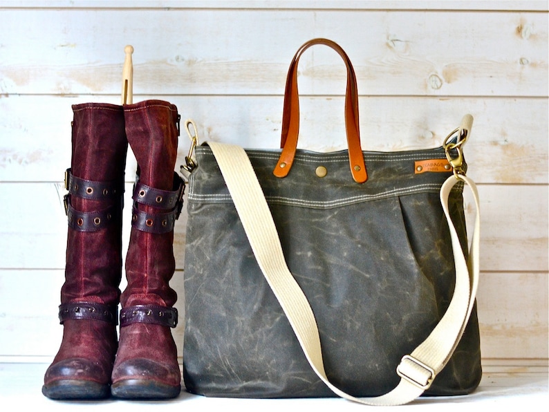 May include: A dark green canvas tote bag with brown leather handles and a white adjustable strap. The bag has a zipper closure and a small pocket on the front. Two burgundy suede boots with buckles are in the background.