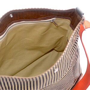 May include: A striped canvas tote bag with a brown leather flap and a zippered pocket. The bag has a bright orange strap.