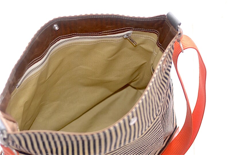 May include: A striped canvas tote bag with a brown leather flap and a zippered pocket. The bag has a bright orange strap.