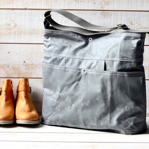May include: A grey waxed canvas messenger bag with a wide adjustable shoulder strap, a top snap closure, and two large front pockets with white stitching, positioned next to a pair of tan leather ankle boots with side buckles and zippers.