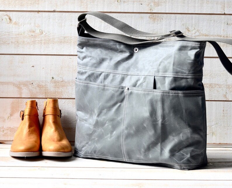 May include: A grey waxed canvas messenger bag with a wide adjustable shoulder strap, a top snap closure, and two large front pockets with white stitching, positioned next to a pair of tan leather ankle boots with side buckles and zippers.