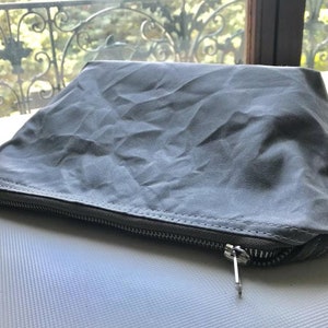 May include: A rectangular gray pouch with a wrinkled, matte texture. The pouch features a horizontal metal zipper closure along the front edge with a silver-toned pull tab. The fabric appears flexible and soft, with visible stitching along the seam.
