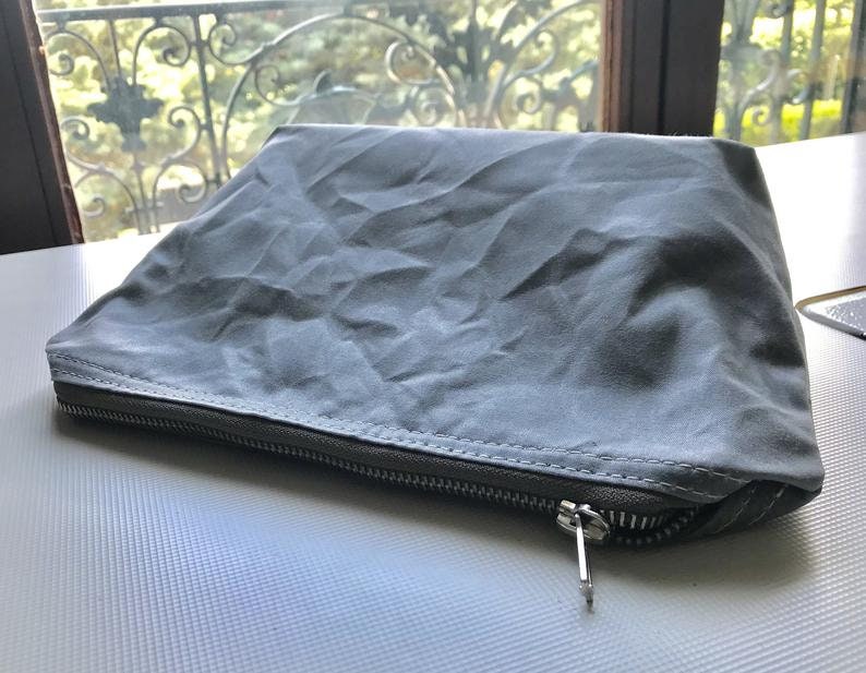 May include: A rectangular gray pouch with a wrinkled, matte texture. The pouch features a horizontal metal zipper closure along the front edge with a silver-toned pull tab. The fabric appears flexible and soft, with visible stitching along the seam.