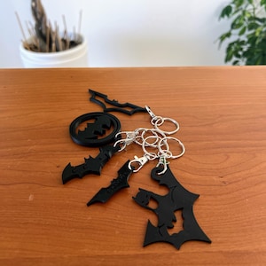 Può includere: Una collezione di portachiavi neri a forma di pipistrello con anelli argentati. I portachiavi presentano vari design, tra cui un simbolo di pipistrello all'interno di un cerchio e sagome di pipistrelli. I portachiavi sono su una superficie di legno.