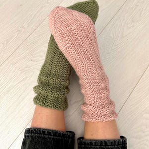 Op de afbeelding: Een paar gebreide sokken, een in lichtroze en de andere in donkergroen. De sokken worden gedragen aan de voeten van een persoon, met de bovenkant van de sokken omgeslagen.