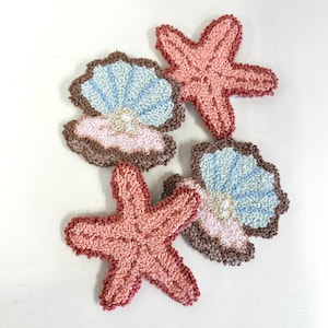 May include: Four handmade, textured coasters in a nautical theme. Two starfish in shades of pink and two seashell designs with blue, pink, and brown accents. The coasters are made of a looped fabric, giving them a unique, handcrafted appearance.