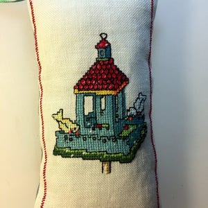 May include: A white fabric pillow with a red embroidered border. The pillow features a cross-stitch design of a blue birdhouse with a red roof and three birds.