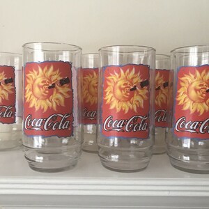 Op de afbeelding: Een set heldere Coca-Cola drinkglazen. Elk glas heeft een rood etiket met een gele zonnegrafiek en het Coca-Cola logo in wit. De glazen zijn cilindrisch met een licht uitlopende basis.