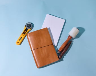 May include: A brown leather bound journal with a black elastic closure, a yellow rotary cutter, a white blank notebook, and two wooden tools with white handles.