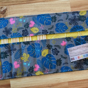 Puede incluir: Un estuche enrollable de tela gris y azul con forro amarillo y una correa negra. El estuche está decorado con un patrón de hojas tropicales y tiene una etiqueta que dice "Epidermium Designs".