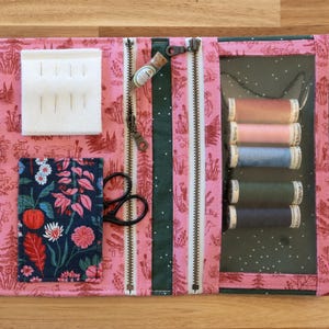 May include: Open sewing kit with a pink floral pattern and green stripes. The kit contains sewing supplies such as thread spools, needles, scissors, and a ruler. The kit is open and laid flat on a wooden surface.