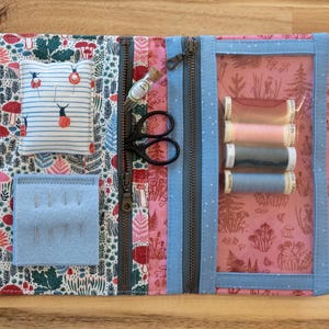 May include: Open sewing organizer with a pink and blue floral pattern. It features clear pockets holding sewing tools like scissors, thread spools, and a needle holder. The organizer is zippered and has a variety of compartments for storage.