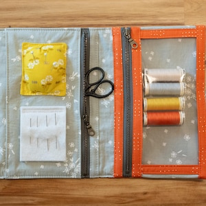 May include: A fabric sewing organizer with light gray floral fabric, orange trim, and clear vinyl pockets. It contains a clear ruler, a white marking pen, a yellow pincushion, a white needle felt pad, black scissors, and four spools of thread.