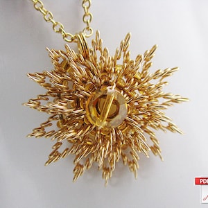 Puede incluir: Un collar con un colgante de oro con un diseño de sol de rayos intrincados. El colgante presenta una piedra redonda de color ámbar en el centro.