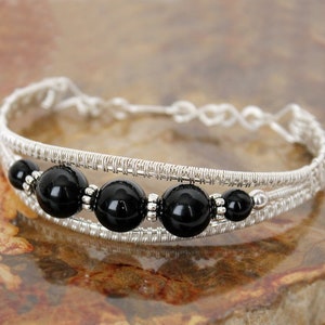 May include: A silver wire bracelet with five large, glossy black beads. The bracelet has a woven design with a double-banded structure and a delicate, openwork closure. The beads are interspersed with smaller silver accents.