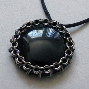 Puede incluir: Un collar con colgante negro con una piedra negra ovalada grande y pulida. La piedra está enmarcada por un borde metálico decorativo con un diseño similar a una cadena. El collar tiene un cordón negro.