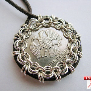 May include: A silver coin pendant featuring a double-headed eagle design, encircled by a chainmail-style border. The coin is set on a dark, circular backing and suspended from a brown cord. The pendant is a unique piece of jewelry.