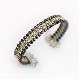 May include: A silver-toned cuff bracelet with a woven design. The bracelet features rows of small, dark brown beads and intricate metalwork. The open-ended design allows for easy wear. The bracelet is displayed on a white background.