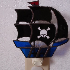 May include: A black and blue stained glass pirate ship night light with a skull and crossbones design. The light is plugged into an outlet.