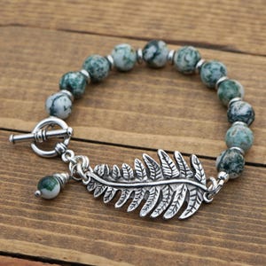 May include: A beaded bracelet featuring round, mottled green and white moss agate stones interspersed with silver-toned metal spacers. The bracelet includes a silver-toned metal toggle clasp and a dangling silver-toned fern leaf charm.