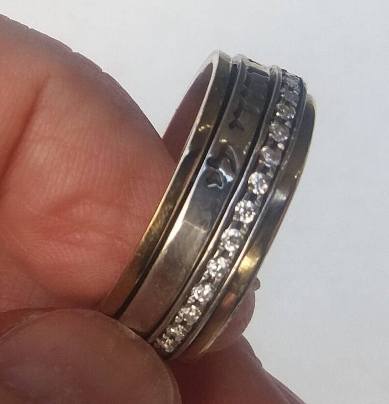K&ouml;nnte beinhalten: Ein drehbarer Ring aus Silber und Gold mit einer Reihe von klaren Zirkonia-Steinen. Der Ring ist mit hebr&auml;ischer Schrift graviert.