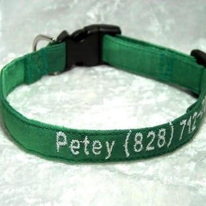 May include: A green dog collar with a black plastic buckle and a silver metal D-ring. The collar is embroidered with the name "Petey" and a phone number in white thread.