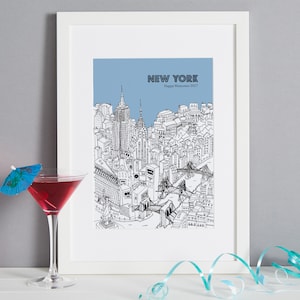 May include: A framed black and white illustration of the New York City skyline, with a light blue sky background. The text "NEW YORK" and "Happy Memories 2017" are printed on the top. A cocktail with a blue umbrella garnish sits in front.