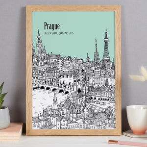 Op de afbeelding: Een zwart-witte lijntekening van de skyline van Praag met de tekst "Praag" en "Jack & Sarah, Kerstmis 2015" onder de afbeelding. De tekening is ingelijst in een licht houten lijst en is geplaatst tegen een lichtgroene achtergrond.