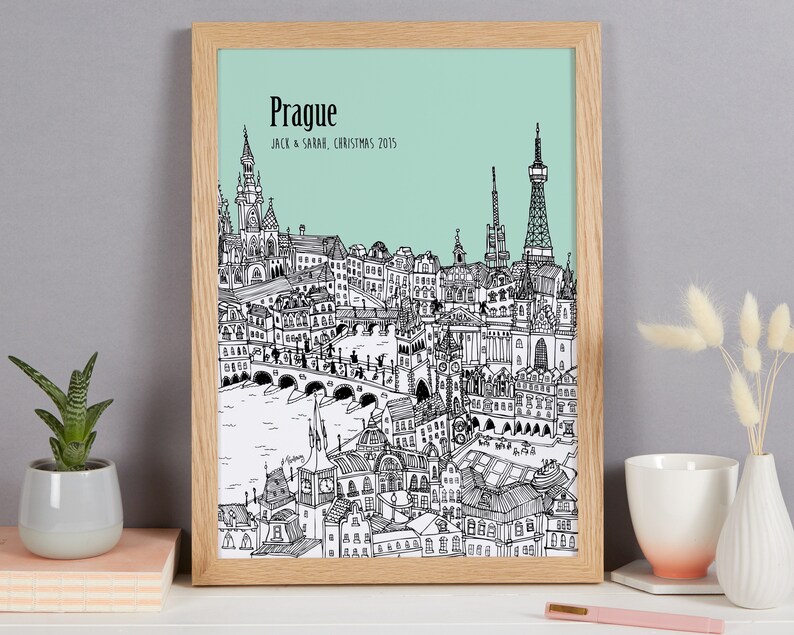 Op de afbeelding: Een zwart-witte lijntekening van de skyline van Praag met de tekst "Praag" en "Jack & Sarah, Kerstmis 2015" onder de afbeelding. De tekening is ingelijst in een licht houten lijst en is geplaatst tegen een lichtgroene achtergrond.