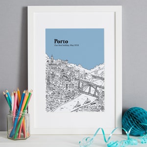 May include: A framed black and white illustration of Porto, Portugal, with a blue sky background. The print includes the city's architecture, a bridge, and the text "Porto, Our first holiday, May 2018". A glass jar of colored pencils and a teal honeycomb ball are in the foreground.
