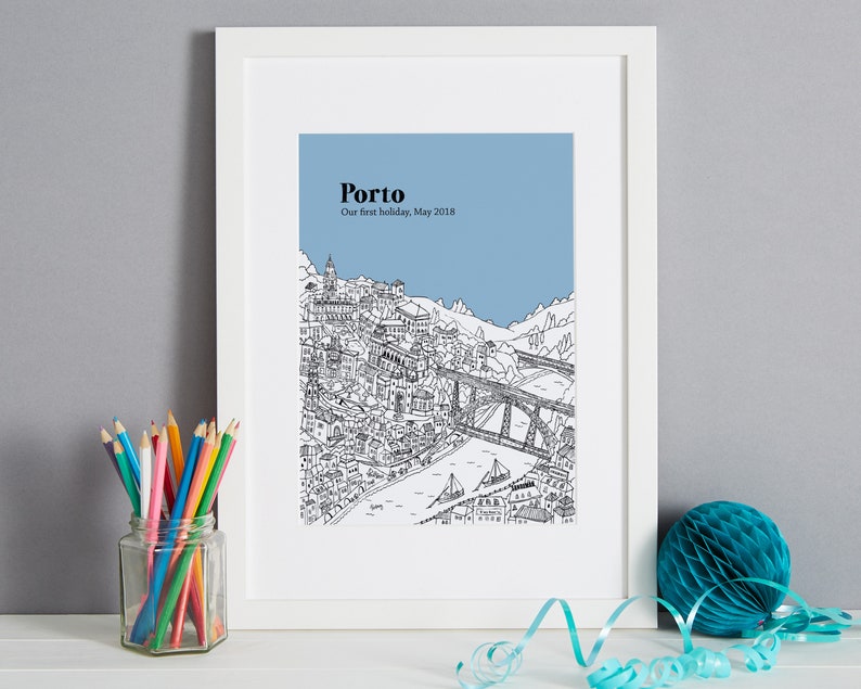 May include: A framed black and white illustration of Porto, Portugal, with a blue sky background. The print includes the city's architecture, a bridge, and the text "Porto, Our first holiday, May 2018". A glass jar of colored pencils and a teal honeycomb ball are in the foreground.