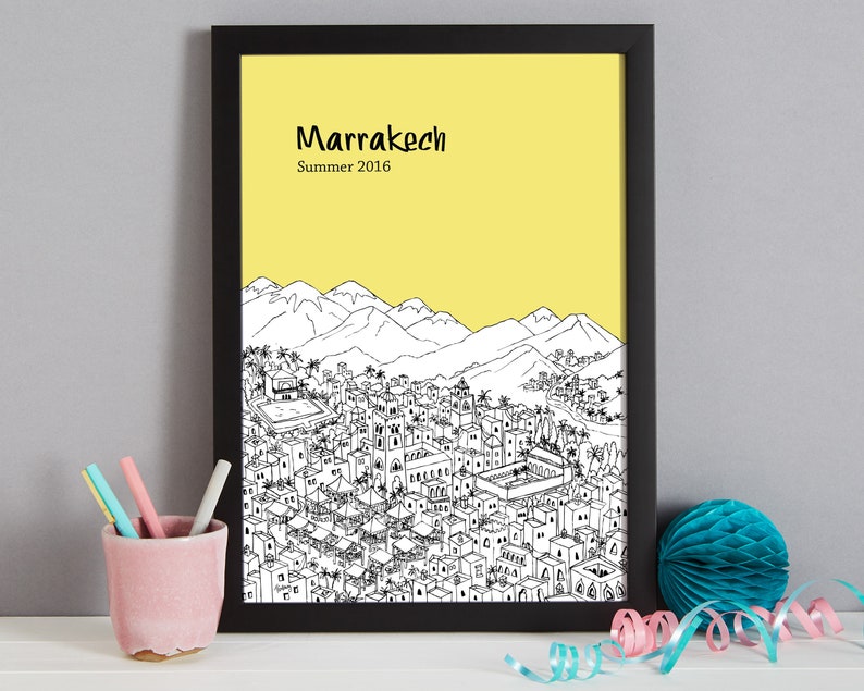 May include: A black framed print with a yellow background and a black and white illustration of the city of Marrakech, Morocco. The text "Marrakech" and "Summer 2016" is printed in black at the top of the print.